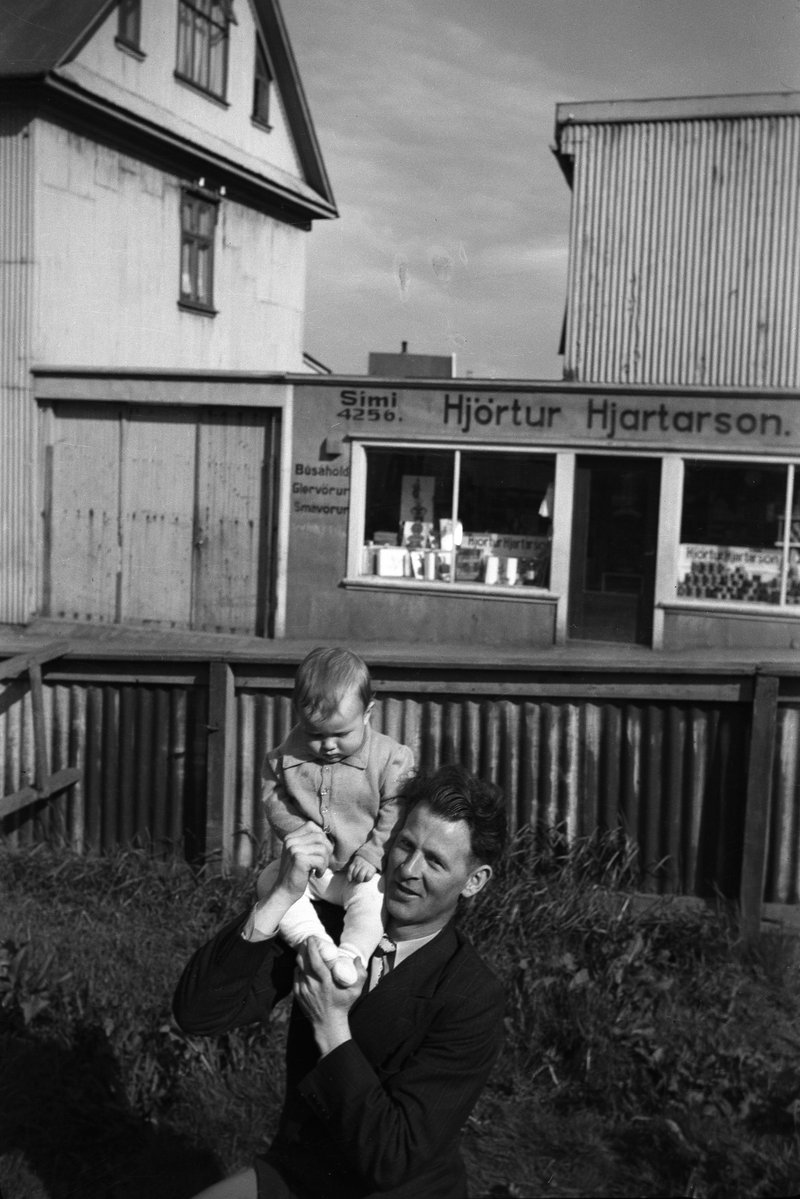 Um 1945-1955, karlmaður með ungt barn á öxlinni. Í baksýn er Bræðraborgarstígur 1, verslun Hjartar Hjartarsonar. Mynd: Kristinn Guðmundsson/Ljósmyndasafn Reykjavíkur