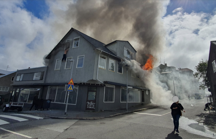 Karlmaður hangir út um glugga á rishæðinni. Hann stökk út þar sem reykur hafði fyllt herbergi hans.