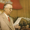 Enver Hoxha í ræðustól með fána albanska kommúnistaflokksins í baksýn.